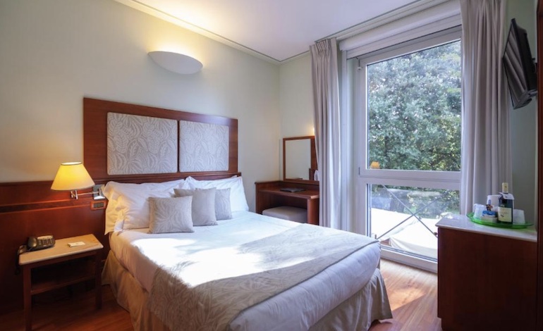 Cama de casal em quarto ensolarado, com janelas e cortinas abertas e vista para jardim. Onde ficar em Lucca.
