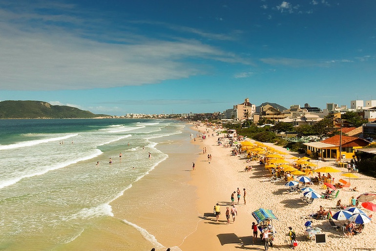 Faixa de areia da Praia dos Ingleses, em dia de céu azul, com muita gente sob o guarda-sol, crianças no mar, vendedores de picolé e pousadas pé na areia.