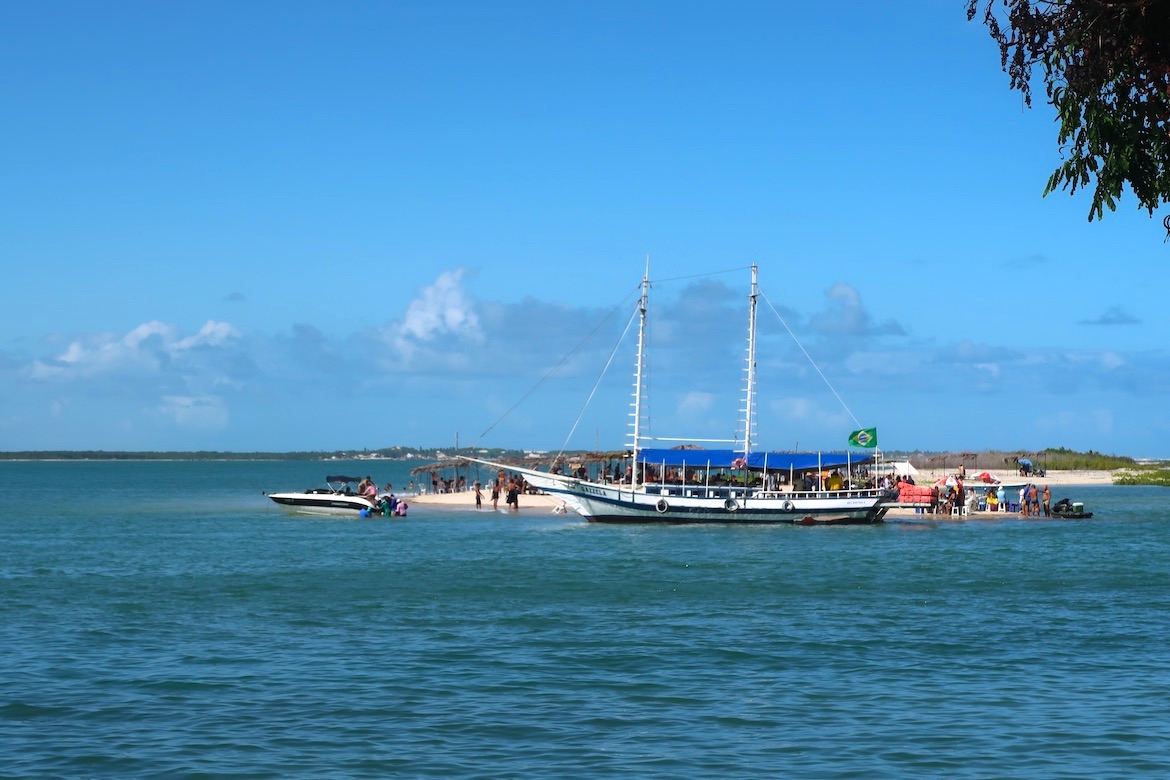 Dia de sol e mar azul em Mangue Seco. Uma escuna com a bandeira do Brasil está ancorada junto à Ilha da Tieta, onde também estão banhistas e um pequeno barco. 